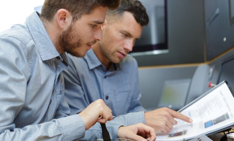Two male technicians look at an equipment manual together. One man points at the handbook while the other looks on.