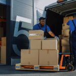 Two logistics employees load a delivery van with cardboard boxes. They're wearing a blue uniform and black hat.