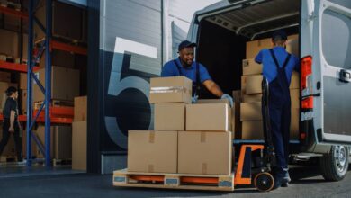 Two logistics employees load a delivery van with cardboard boxes. They're wearing a blue uniform and black hat.