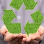 A person wearing a gray button down shirt holds out their hands to cradle a large green recycling symbol.