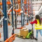 A woman wearing a hi-visibility yellow vest pushes a pallet jack into an aisle between two orange and blue pallet racks.