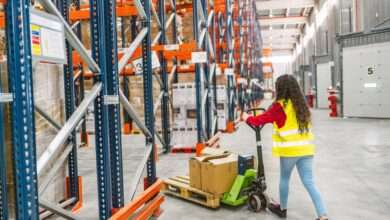 A woman wearing a hi-visibility yellow vest pushes a pallet jack into an aisle between two orange and blue pallet racks.