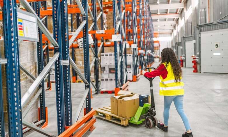 A woman wearing a hi-visibility yellow vest pushes a pallet jack into an aisle between two orange and blue pallet racks.