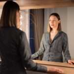 A woman wearing a black and white striped shirt stands in front of a well-lit rectangular mirror.