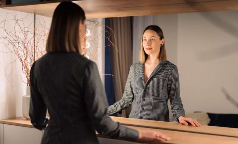 A woman wearing a black and white striped shirt stands in front of a well-lit rectangular mirror.