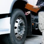 A man in cargo pants holds a pen and clipboard as he touches the wheel of a semitruck to inspect it.