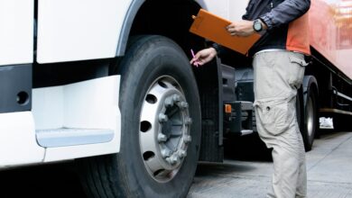 A man in cargo pants holds a pen and clipboard as he touches the wheel of a semitruck to inspect it.