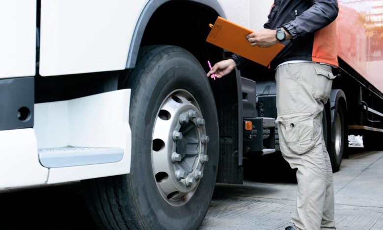 A man in cargo pants holds a pen and clipboard as he touches the wheel of a semitruck to inspect it.