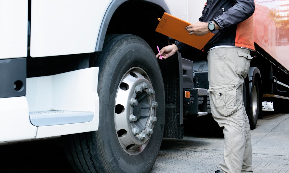 A man in cargo pants holds a pen and clipboard as he touches the wheel of a semitruck to inspect it.