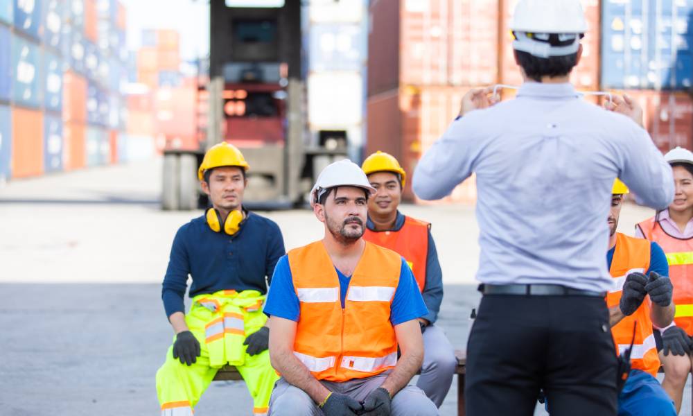 A group of five shipyard workers in PPE sit and pay attention to a man facing them to give instructions.