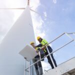 Two men wearing hard hats and safety vests look at a blueprint while standing next to a giant wind turbine.