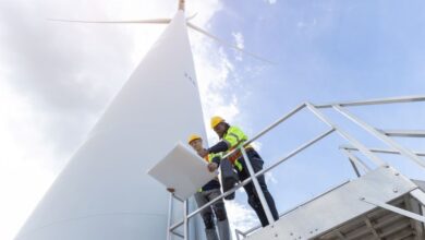 Two men wearing hard hats and safety vests look at a blueprint while standing next to a giant wind turbine.