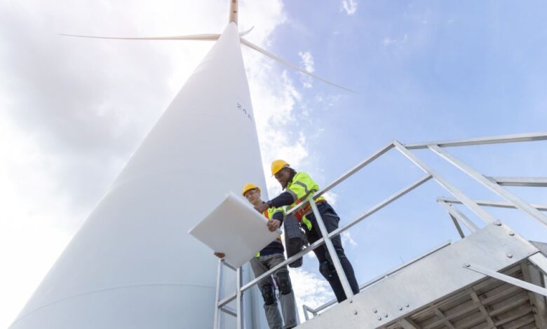 Two men wearing hard hats and safety vests look at a blueprint while standing next to a giant wind turbine.
