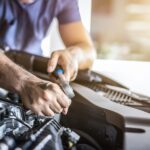 A close-up of a man in a blue T-shirt who is using a small tool to work on the engine of a vehicle in a garage.