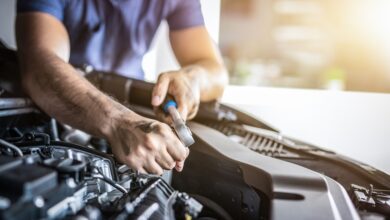 A close-up of a man in a blue T-shirt who is using a small tool to work on the engine of a vehicle in a garage.
