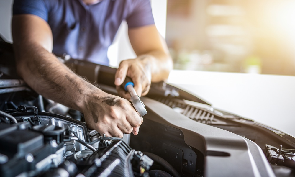 A close-up of a man in a blue T-shirt who is using a small tool to work on the engine of a vehicle in a garage.
