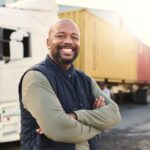 A smiling, industrial Black male truck driver is standing in front of a large semi-truck used to transport goods.