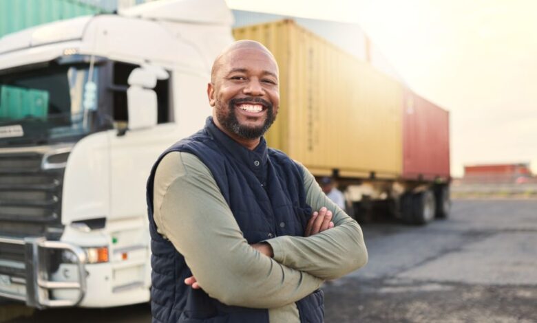 A smiling, industrial Black male truck driver is standing in front of a large semi-truck used to transport goods.