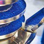 Small blue granules of plastic moving through a metal dispenser as part of a manufacturing process.