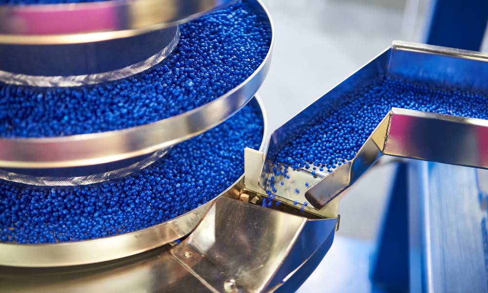 Small blue granules of plastic moving through a metal dispenser as part of a manufacturing process.