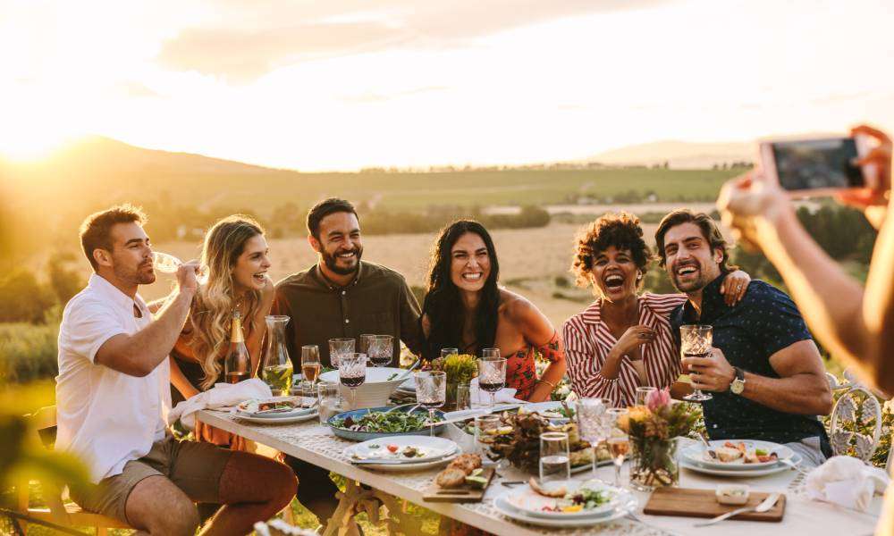 A person taking a photo of different people sitting at a large table outdoors. They are all smiling and laughing together.