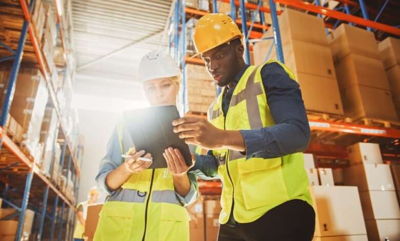 Two warehouse workers in safety vests and hard hats look closely at an electronic tablet in front of boxes on shelves.