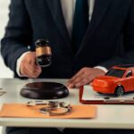 A man wearing a black suit sits at a desk with a gavel, clipboard, red model car, pen, and various items in front of him.