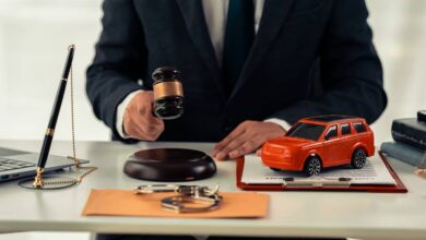 A man wearing a black suit sits at a desk with a gavel, clipboard, red model car, pen, and various items in front of him.