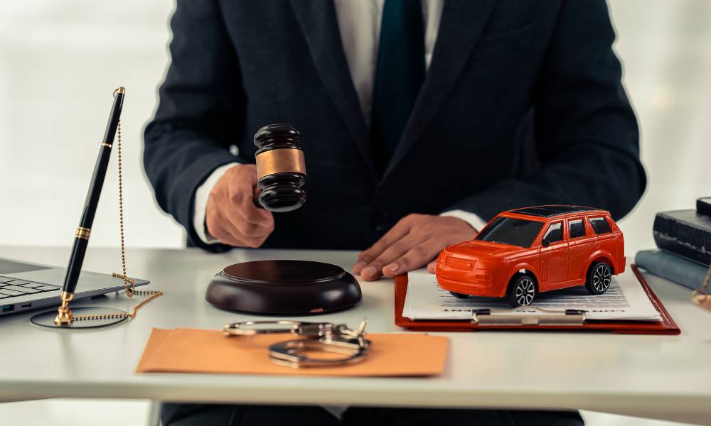 A man wearing a black suit sits at a desk with a gavel, clipboard, red model car, pen, and various items in front of him.