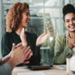 In a business meeting, employees around a table clap for a woman smiling and holding her hands to her chest.