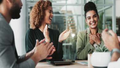 In a business meeting, employees around a table clap for a woman smiling and holding her hands to her chest.