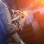 A man in a gray suit holding a microphone and a pad of paper with orange and yellow lights in the background.