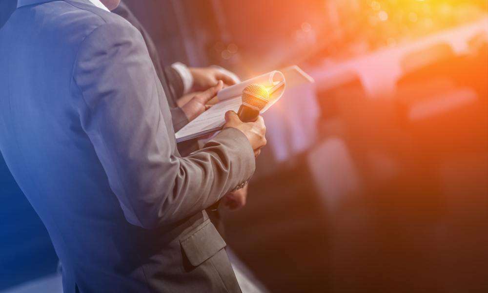 A man in a gray suit holding a microphone and a pad of paper with orange and yellow lights in the background.