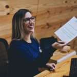 A grinning receptionist sits at her desk and looks up at the person she's handing a piece of paper to.