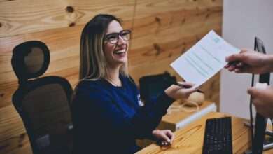 A grinning receptionist sits at her desk and looks up at the person she's handing a piece of paper to.