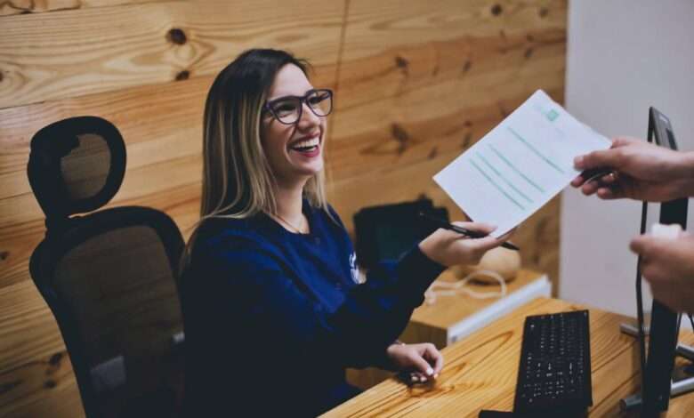 A grinning receptionist sits at her desk and looks up at the person she's handing a piece of paper to.
