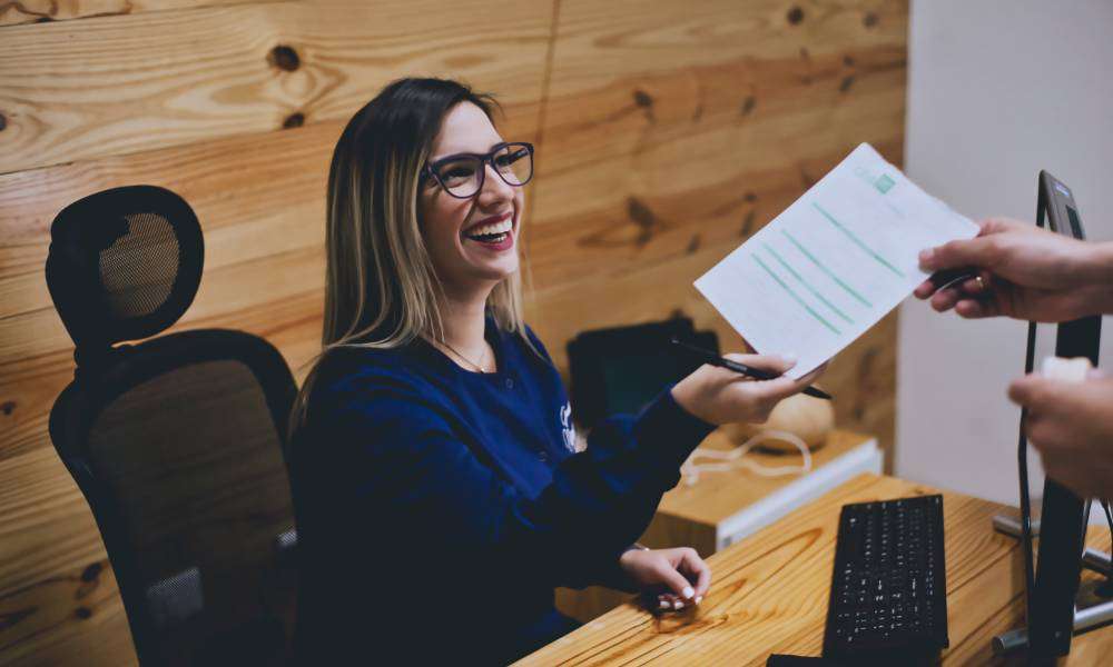 A grinning receptionist sits at her desk and looks up at the person she's handing a piece of paper to.