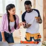 A Man and a woman standing inside a room undergoing renovations with tools on a desk, looking at a smartphone.