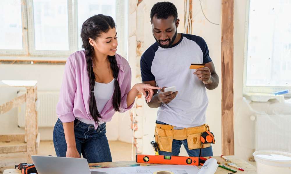 A Man and a woman standing inside a room undergoing renovations with tools on a desk, looking at a smartphone.