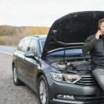A man sitting on his parked car at the edge of a highway. He is on the phone with his hood propped open.