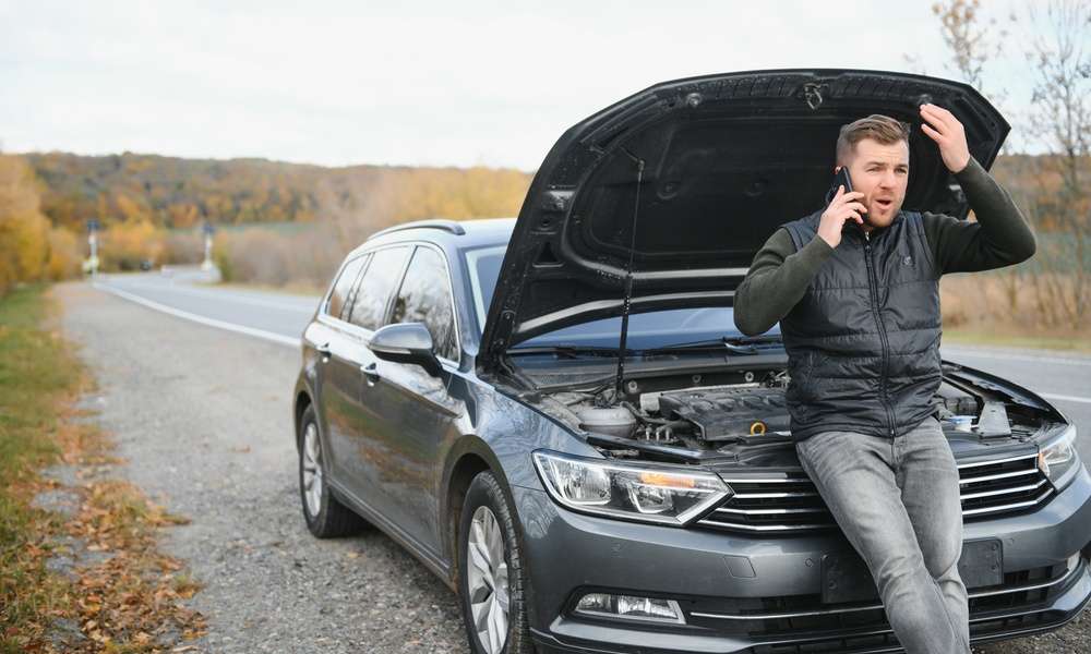A man sitting on his parked car at the edge of a highway. He is on the phone with his hood propped open.