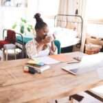 A woman working remotely at a home desk, reaching for her laptop that's next to planners, markers, and digital devices.