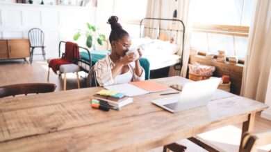 A woman working remotely at a home desk, reaching for her laptop that's next to planners, markers, and digital devices.