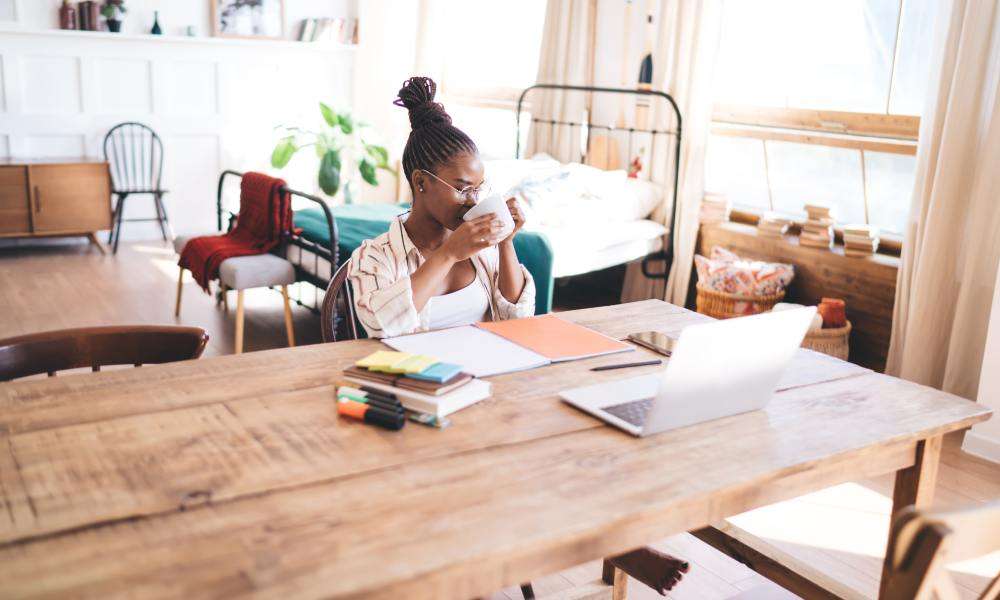 A woman working remotely at a home desk, reaching for her laptop that's next to planners, markers, and digital devices.