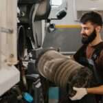 An auto mechanic wearing black overalls and white work gloves, taking out a dirty air filter from a fleet truck.