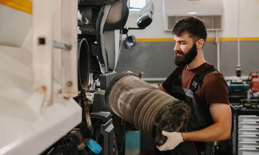 An auto mechanic wearing black overalls and white work gloves, taking out a dirty air filter from a fleet truck.