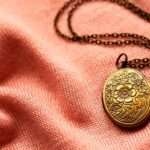 An antique bronze necklace lying against a pink fabric. There is a flower design on the necklace with a long chain.