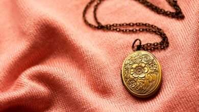 An antique bronze necklace lying against a pink fabric. There is a flower design on the necklace with a long chain.