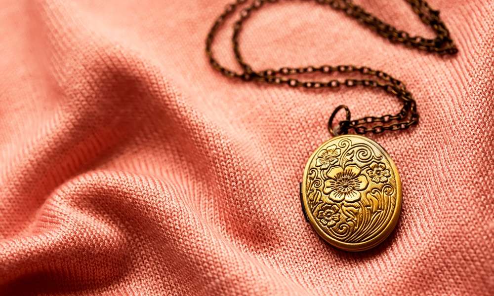 An antique bronze necklace lying against a pink fabric. There is a flower design on the necklace with a long chain.