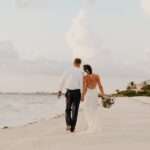A bride and groom walk away on the beach as they hold hands. They're leaning in toward one another.